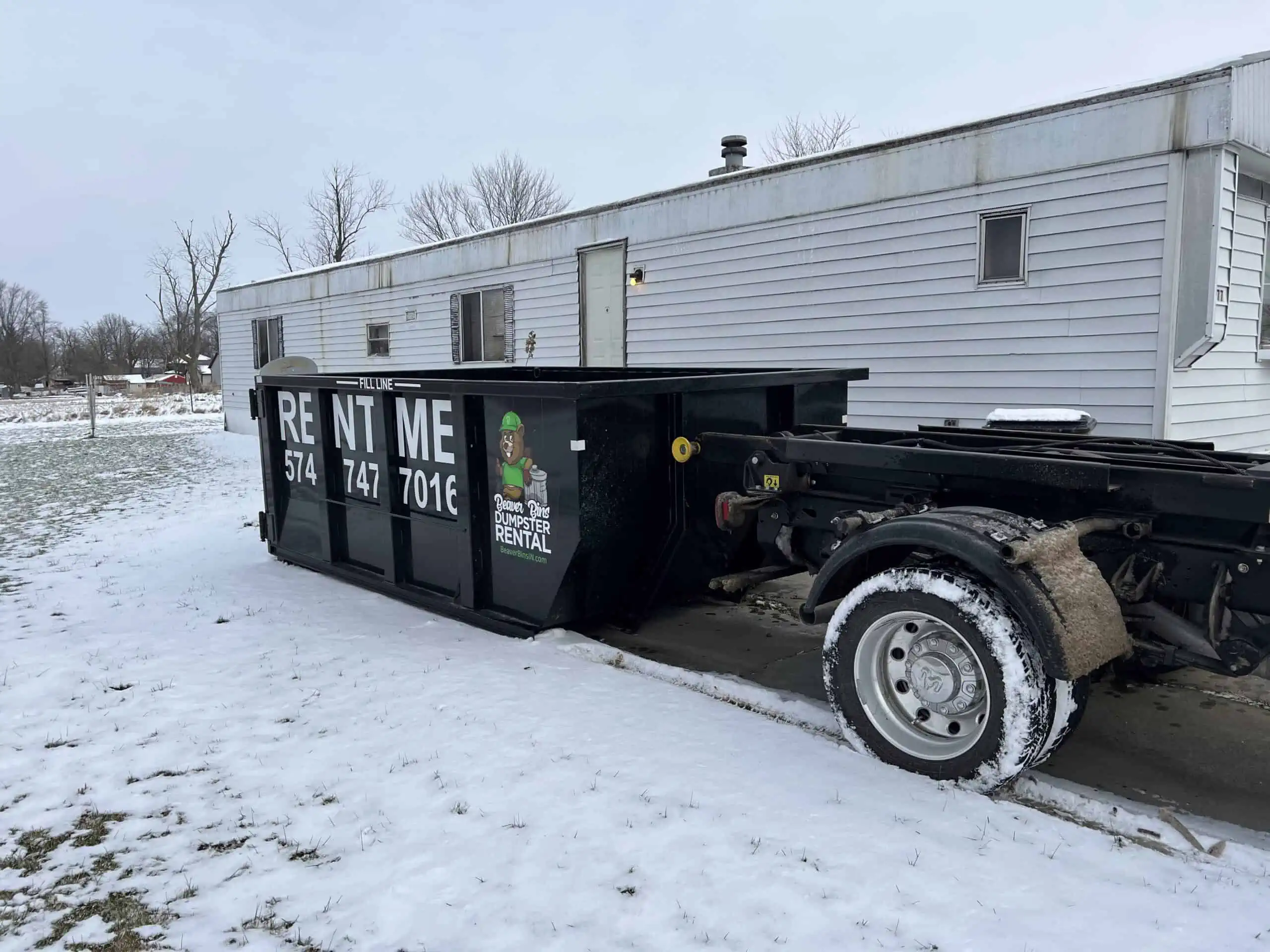 a black Beaver Bins dumpster hooked up to a trailer in the snow outside a mobile home
